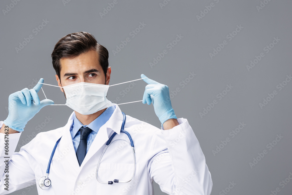 A male doctor in a white coat and medical mask and sterile gloves looks at the camera on a gray isolated background, copy space, space for text A male doctor in a white coat and medical mask and sterile gloves looks at the camera on a gray isolated background, copy space, space for text