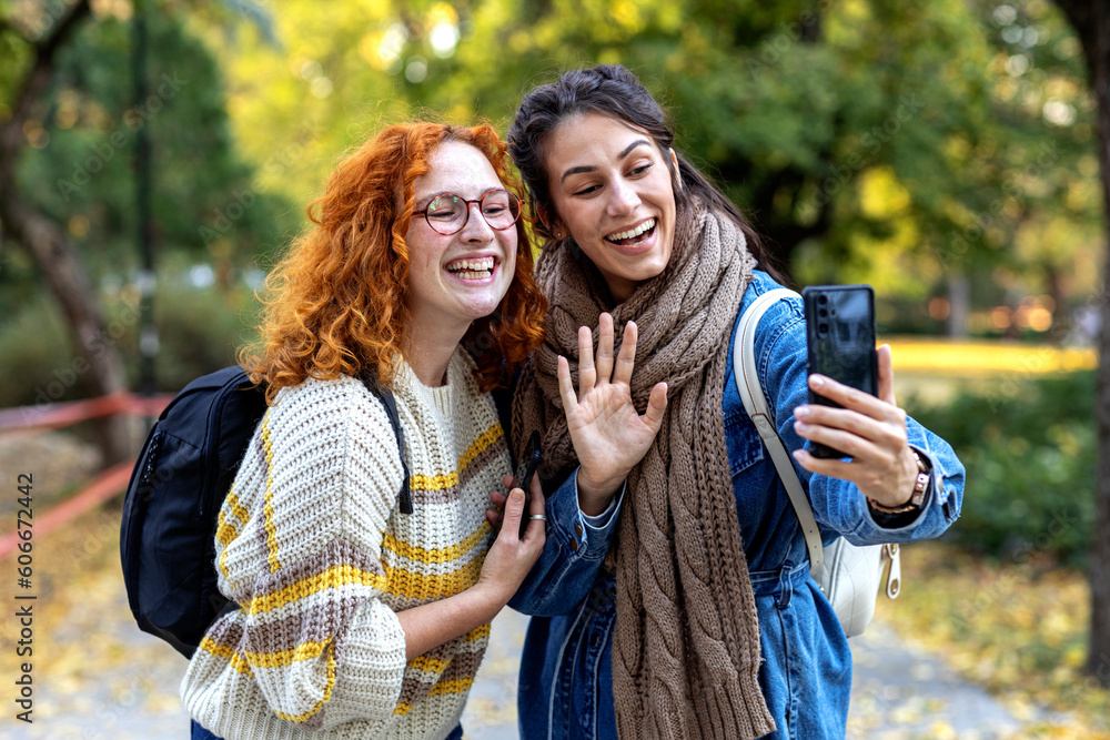 Fototapeta premium Two student woman friends in urban public park use mobile phone and smile