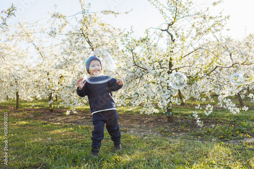 A little boy, 3 years old, in a sweater and a hat, runs through a blooming garden. Clothes for children aged 3 years. A happy eotian child is white among blossoming apple trees © Дмитрий Ткачук