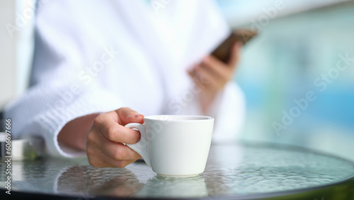 Woman in bathrobe with mobile phone in hands drinking coffee from white cup at table in spa closeup. Rest with cup of fragrant tea concept