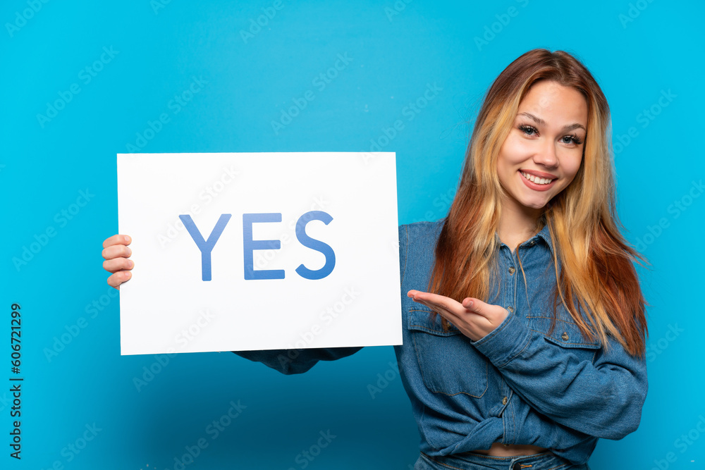 Teenager girl over isolated blue background holding a placard with text YES and pointing it Teenager girl over isolated blue background holding a placard with text YES and pointing it