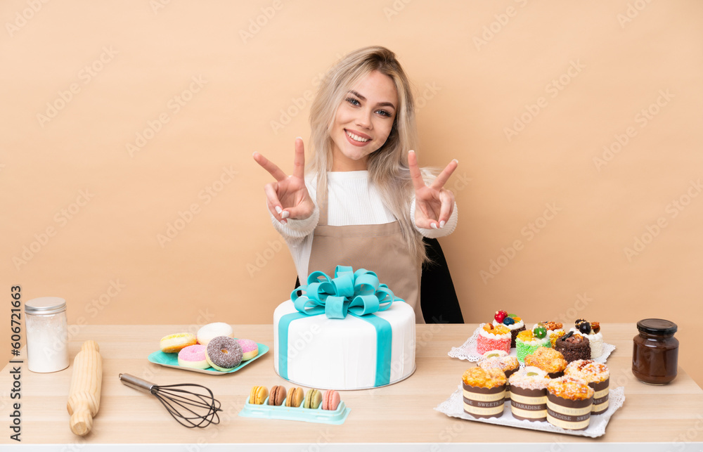 Teenager pastry chef with a big cake in a table smiling and showing victory sign Teenager pastry chef with a big cake in a table smiling and showing victory sign