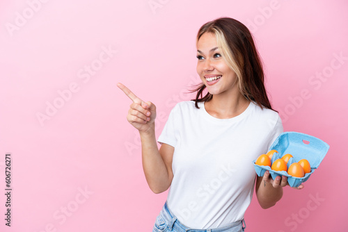 Young Russian woman holding eggs isolated on pink background pointing finger to the side and presenting a product