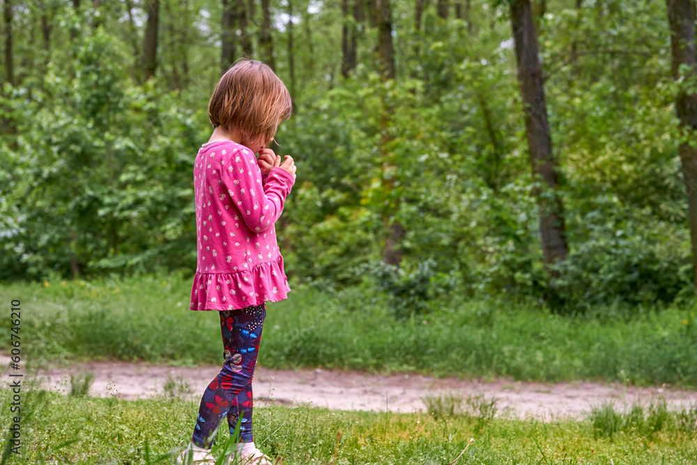 A little preschool child girl in a pink dress walks in a green forest, thinking