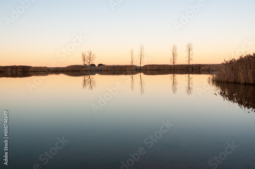 Vista de un lago en calma al atardecer con árboles y su reflejo en el agua en calma.