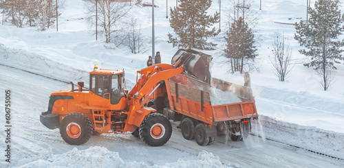 Big orange tractor cleans up snow from the road and loads it into the truck. Cleaning and cleaning of roads in the city from snow in winter. Snow removal after snowfall and blizzards. 