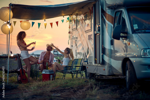 Three friends toasting and having fun outdoors, in front of camper rv. Summertime sunset. Travel, holiday, weekend, togetherness, lifestyle concept.