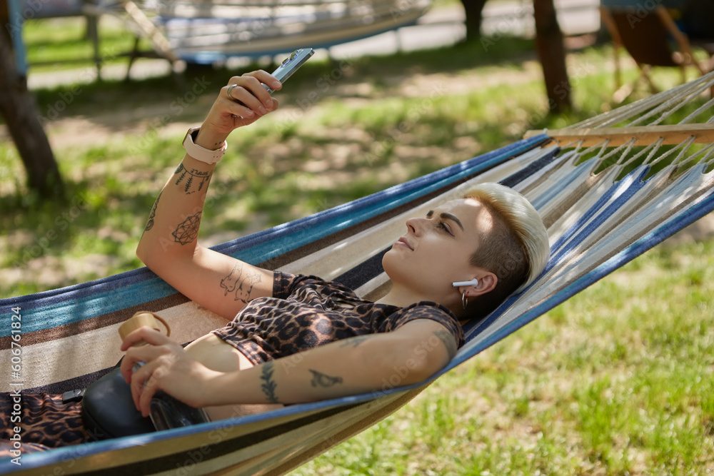 Stylish young woman with short dyed hair lying on a hammock in a sunny green park, listening to music and browsing internet on a smart phone. Diverse female person relaxing outdoor on weekend Stylish young woman with short dyed hair lying on a hammock in a sunny green park, listening to music and browsing internet on a smart phone. Diverse female person relaxing outdoor on weekend