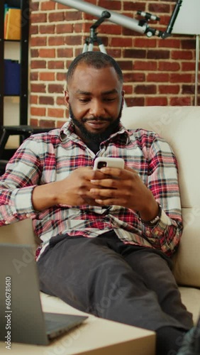 Cozy african american man chatting with online friends over the phone in warm stylish home, enjoying himself. Happy smiling person scrolling on phone entertained by social media activity