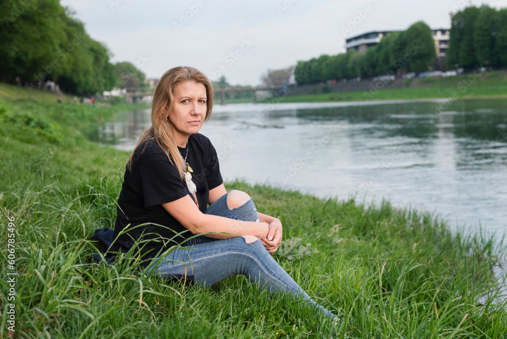 Φωτογραφία Confused, disappointed middle-aged woman sitting on grass in city in park area near river, woman offended at man
