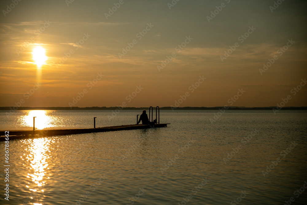 the silhouette of a woman sitting on a wooden jetty by the water