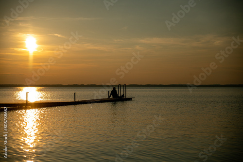 the silhouette of a woman sitting on a wooden jetty by the water