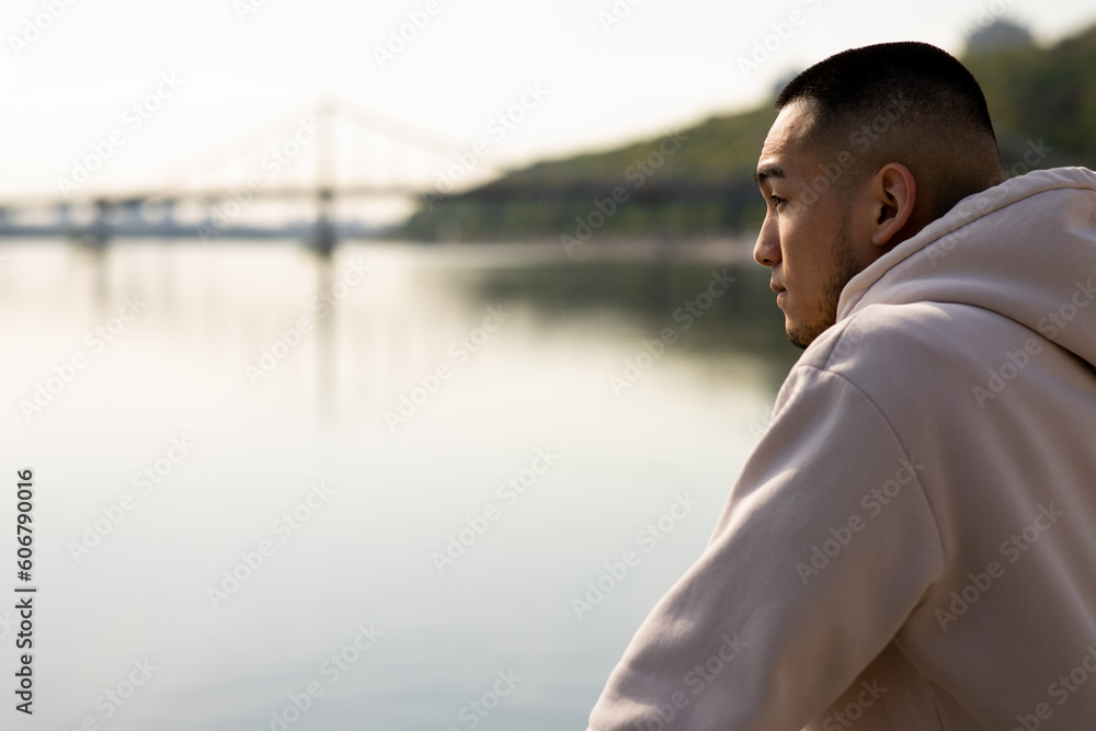 A man with a short haircut stands near the river and looks into the distance. A young Asian guy in a hoodie stands against the backdrop of a blurred river.