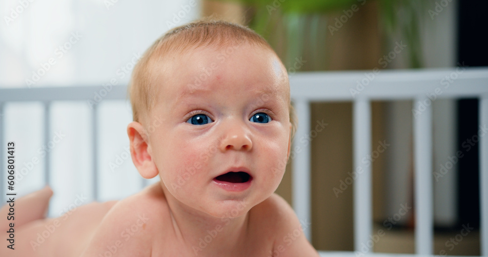 Close up. Portrait of beautiful infant kid is lying on the white bed on his stomach. A nice little caucasian newborn baby with blue eyes.