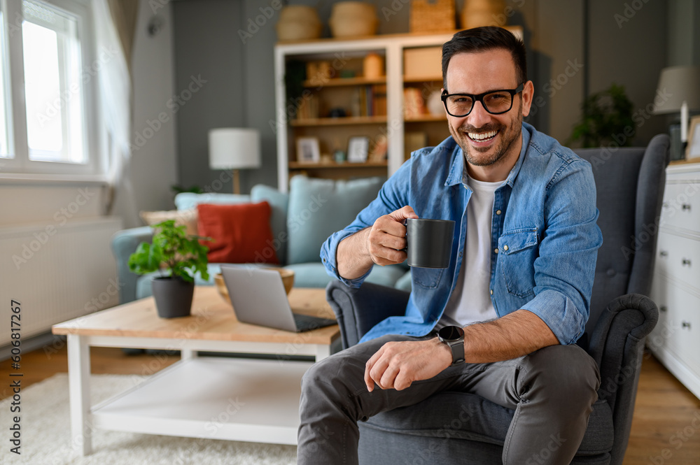 Portrait of confident male entrepreneur holding coffee cup and smiling cheerfully while sitting on armchair. Young happy professional taking coffee break while working in home office