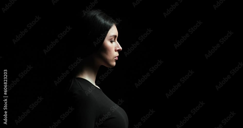 Woman stands in profile against a black background. She slowly and dramatically lifts her head upwards and gazes at the light with a serious and thoughtful expression. Woman stands in profile against a black background. She slowly and dramatically lifts her head upwards and gazes at the light with a serious and thoughtful expression.