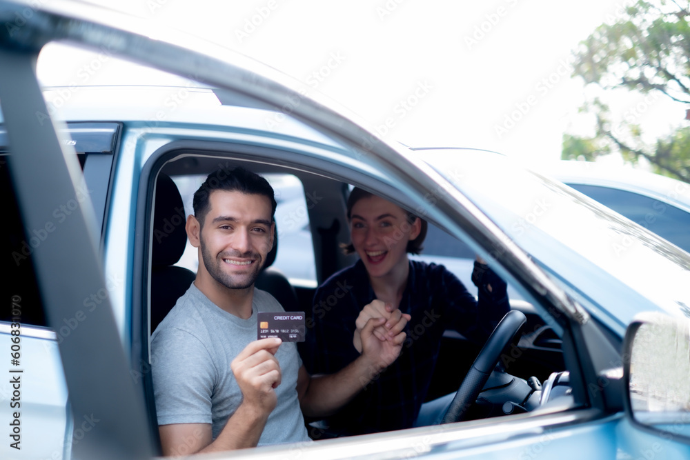 White handsome man holding credit card looking to camera with smile while holding his happy girlfriend hand sitting in a car, using financial product concept.