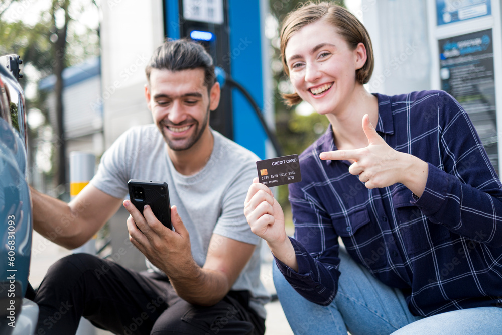 Young handsome Arub man looking on his smartphone with smile of happiness, easy pay for charging service for electronic car green power while her girl friend point and show credit card.
