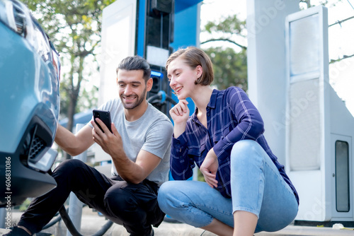 Couple of white persons male and female sitting low in front of Electrical car energy station  looing at smart phone with teeth smile. photo