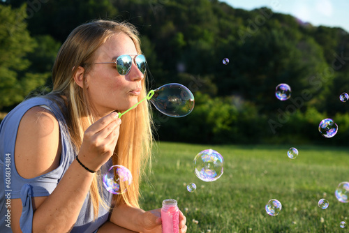 Blonde woman with aviator sunglasses blowing bubbles at golden hour on a warm summer evening.