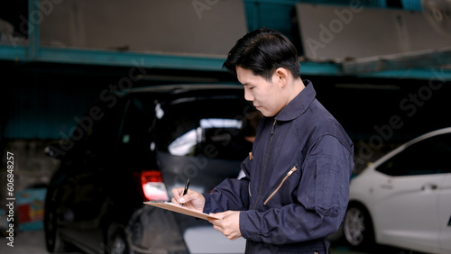 Asian insurance worker standing in mechanic suit Standing holding documents to inspect the condition of car that has been involved in accident To prepare to estimate cost of repairing the car.