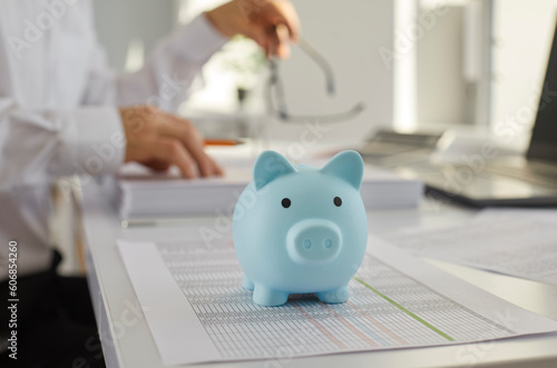 Blue piggy bank on the table with a businessman accountant in background working at home or at office with documents, tables and charts making calculations. Investment and savings concept.