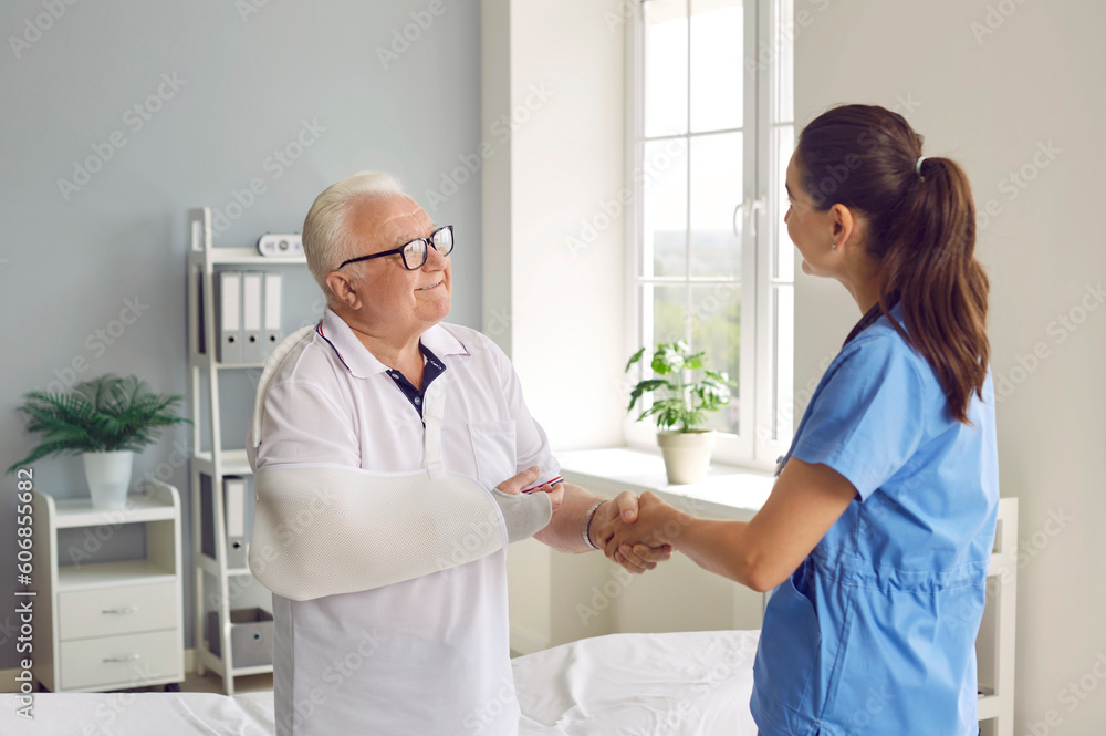 Fototapeta premium Happy, grateful male patient exchanging handshakes with doctor. Senior man with broken arm shaking hands with young female surgeon, thanking her for help and smiling. Injury treatment concept