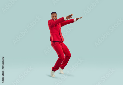 Cheerful excited stylish african american man having fun on pastel gray background. Happy fashionable man in red suit dances funny standing on his toes with outstretched arms. Full length.