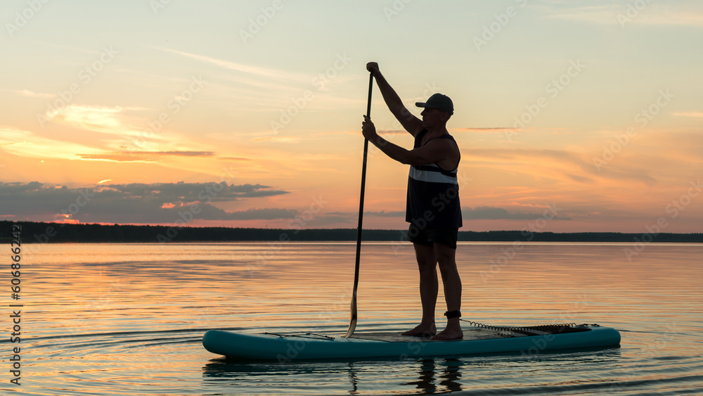A man on a SUP board with a paddle at sunset against a pink-orange sky swims in the water of the lake.