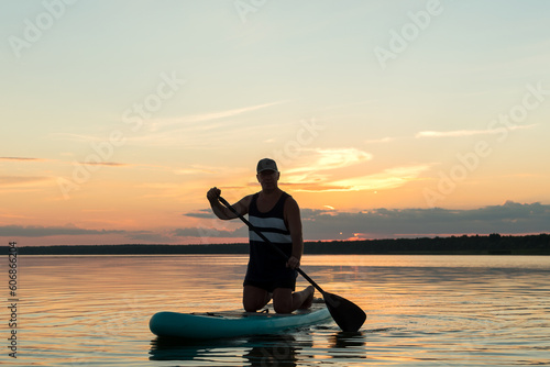 A man on his knees on a SUP board with an oar swims in the calm water of the lake against the backdrop of a pink sky reflection in the water. © finist_4