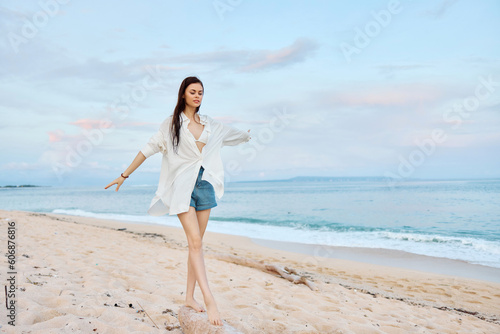 Happy tanned woman in white swimsuit shirt and denim shorts walks on the beach on the sand by the ocean with wet hair after swimming  sunset light and pink clouds in Bali
