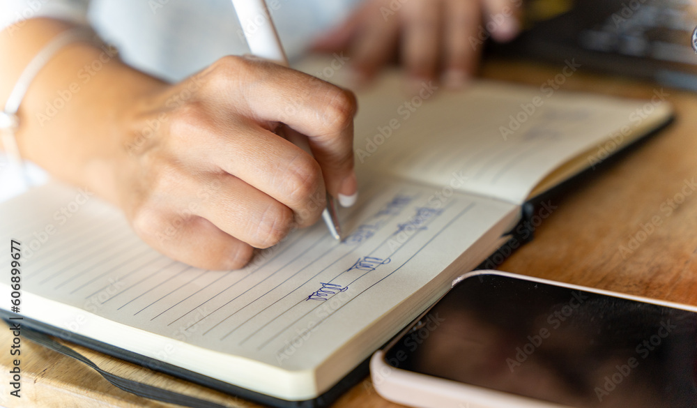 Close-up view of a woman writing on a notebook while working with a laptop in a coffee shop. Close-up view of a woman writing on a notebook while working with a laptop in a coffee shop.