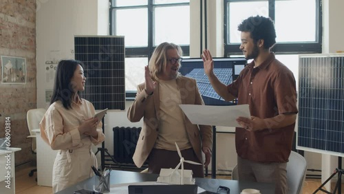 Medium full shot of three multiethnic male and female architects discussing final plan of zero carbon residential property with renewable energy sources, then smiling, cheering and doing high fives photo