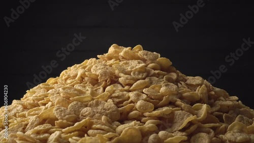 A pile of yellow corn flakes slowly rotates on a dark black background. Concept of healthy food - cereal breakfast. Close-up, side view. Copy space for advertising text