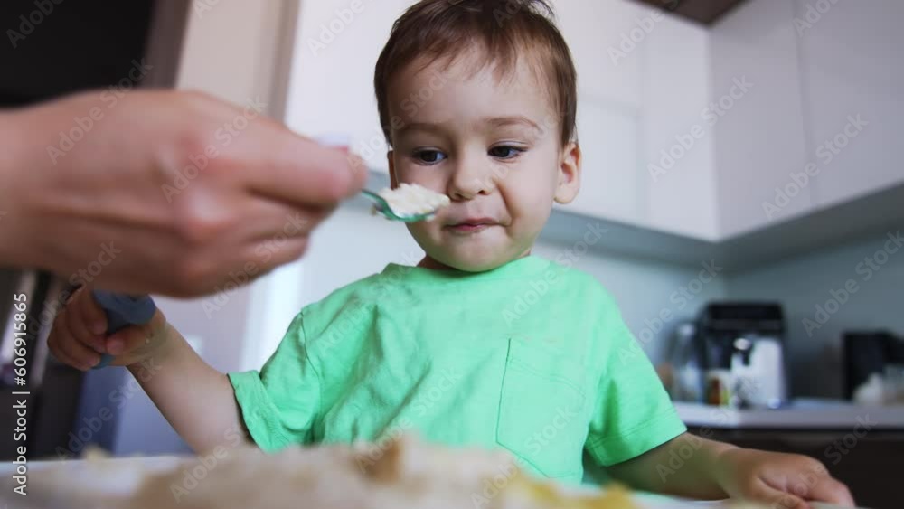 Adorable toddler having meals. Kid holds the fork but eats from mom's spoon. Low angle view. Adorable toddler having meals. Kid holds the fork but eats from mom's spoon. Low angle view.