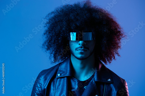 Portrait of fashion man with curly hair with stylish glasses on blue background multinational, colored light, black leather jacket trend, modern concept.
