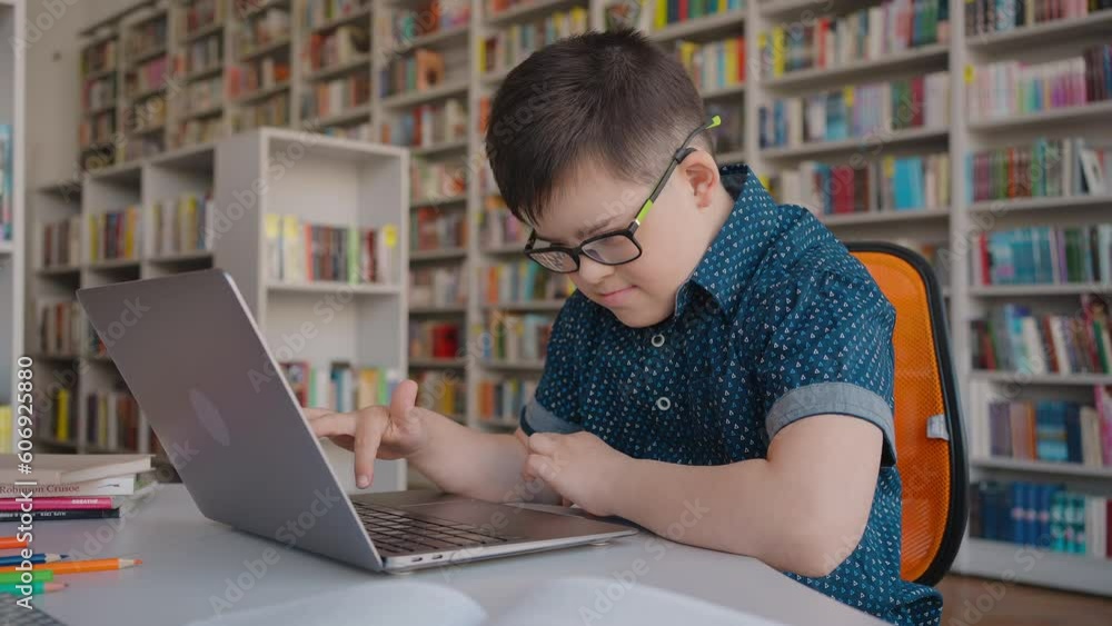 Adorable school child boy Down syndrome typing text on laptop, studying online in library. Adorable school child boy Down syndrome typing text on laptop, studying online in library.