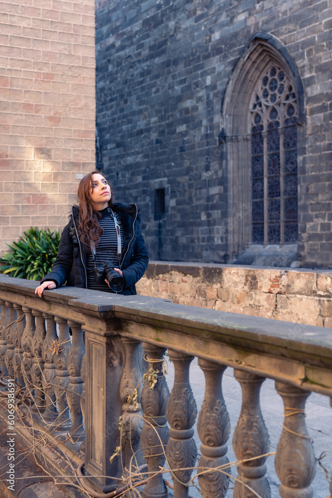 Thoughtful young woman tourist with camera admiring old Barcelona Cathedral in Gothic Quarter while standing against brick wall during sightseeing trip in Spain