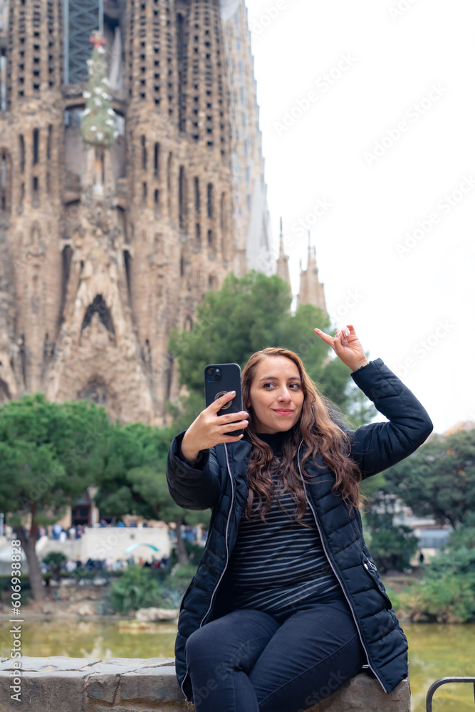 Serious young Latin woman traveler in warm outfit with photo camera rest on a park bench against Sagrada Familia by Antonio Gaudi during sightseeing tour in Barcelona, Spain