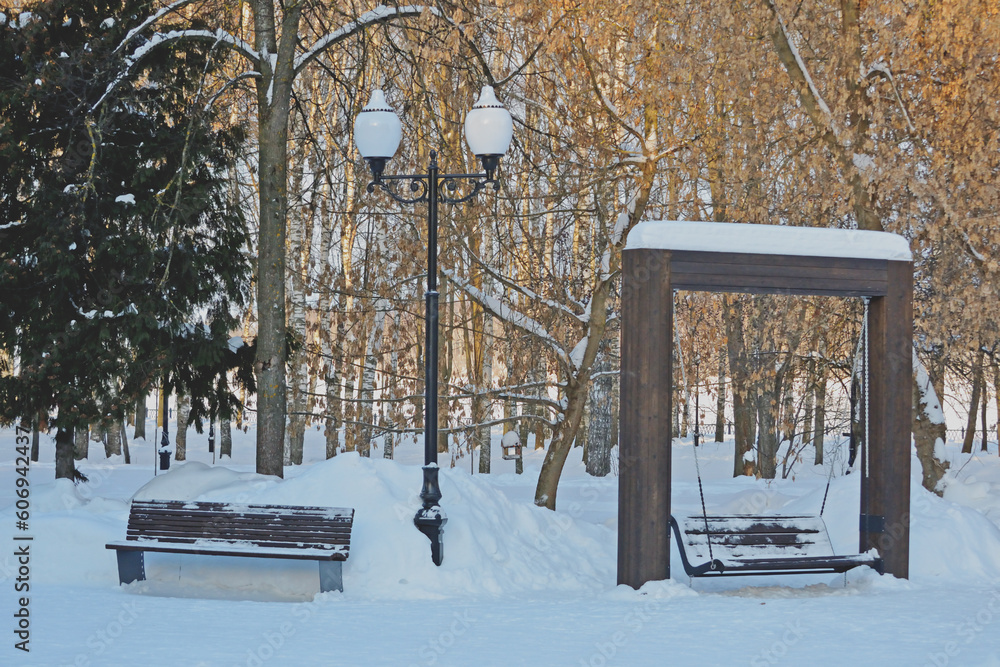 Φωτογραφία Snowdrifts, snow-covered trees, benches and swings in a city park in winter at sunset