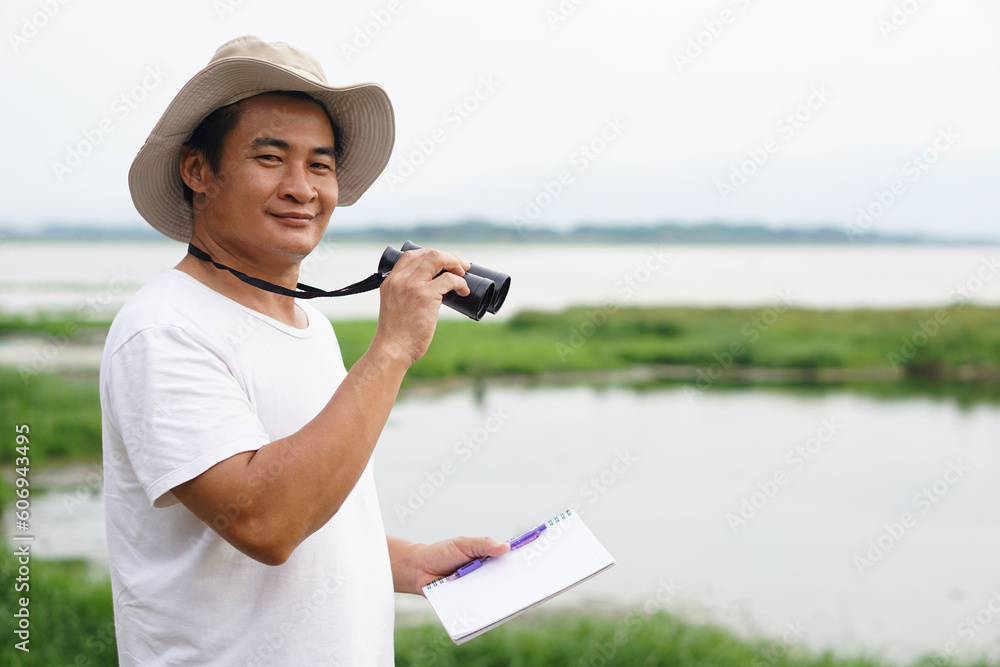 Handsome Asian man ecologist is surveying nature at the lake, holds binoculars and paper notebook. Concept, nature exploration. Ecology study.  Pastime activity, lifestyle. Man explore environment    