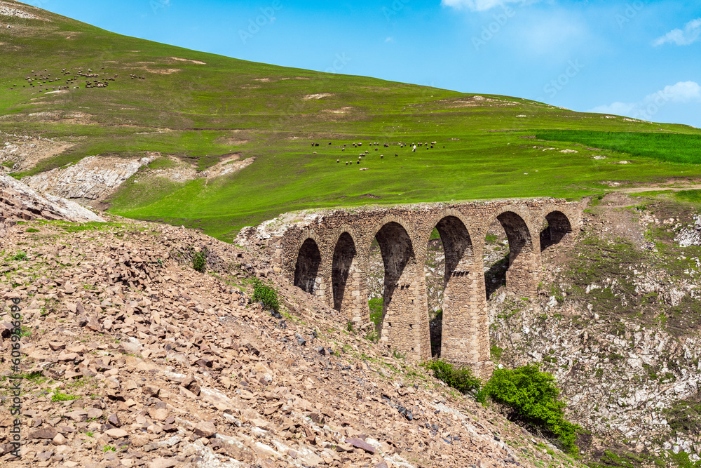 A flock of sheep grazes on a green mountain slope near an ancient stone bridge