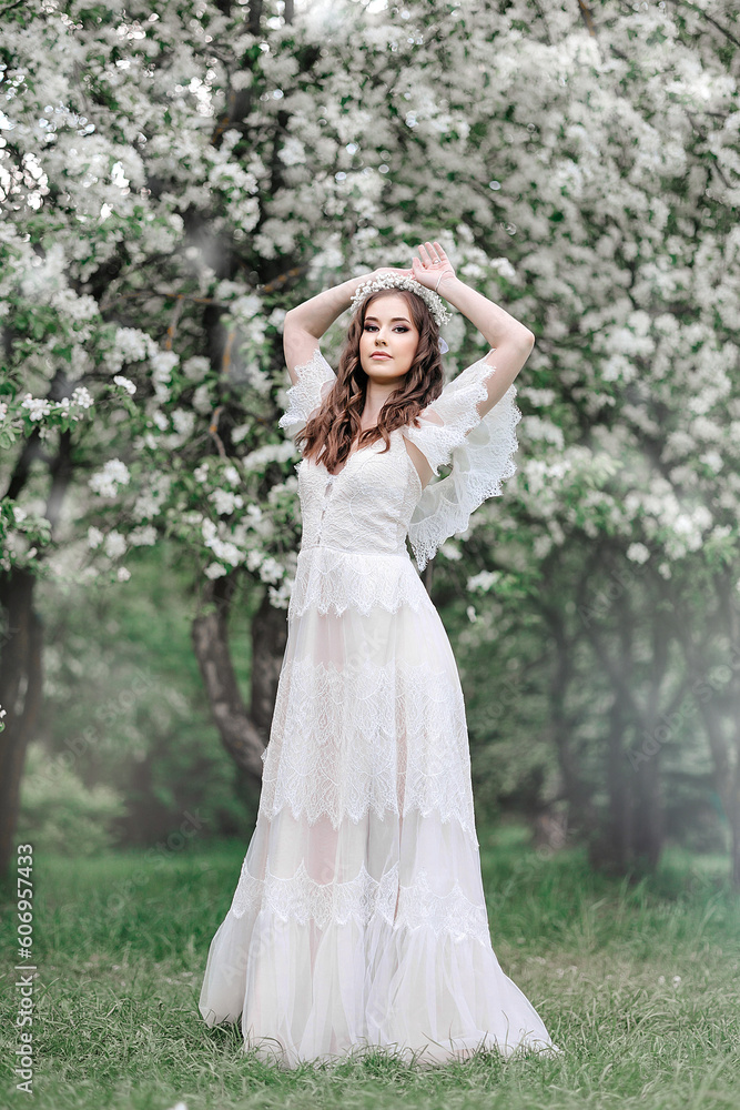 A beautiful young girl in a white floor-length lace dress in a blooming apple orchard