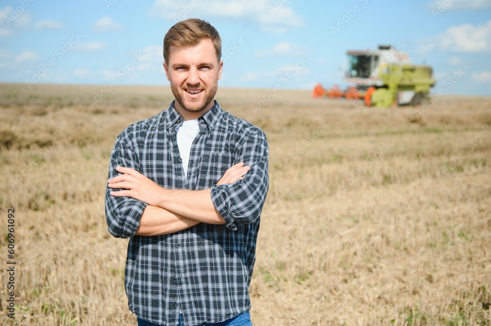 Happy farmer proudly standing in a field. Combine harvester driver going to crop rich wheat harvest. Agronomist wearing flannel shirt, looking at camera on a farmland
