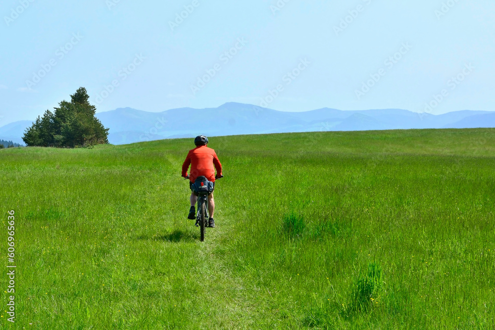 Back view of elderly man in helmet riding bicycle on grass hill. Misty mountains in the background