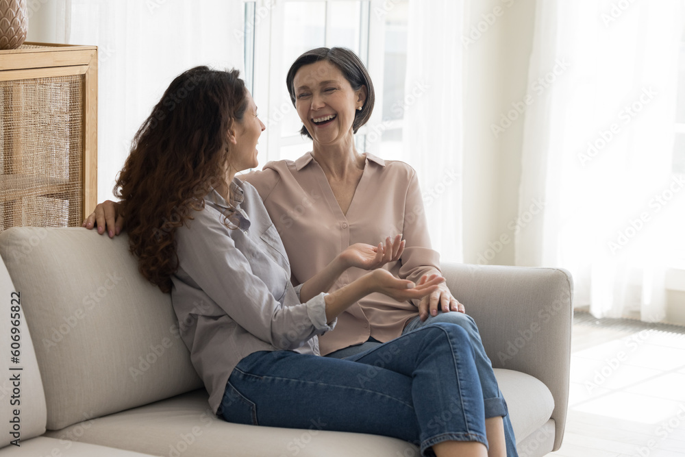 Cheerful senior mature pretty mom and adult daughter woman meeting at home, talking, discussing good news, family gossip, sitting close, speaking, laughing with joy