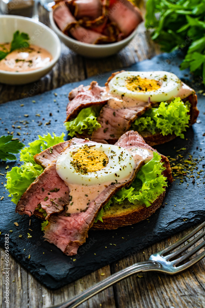 Delicious sandwiches with grilled beef sirloin hard boiled eggs and lettuce on wooden table

