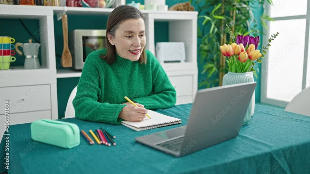 Mature hispanic woman with grey hair sitting on table having online painting lesson at dinning room Mature hispanic woman with grey hair sitting on table having online painting lesson at dinning room