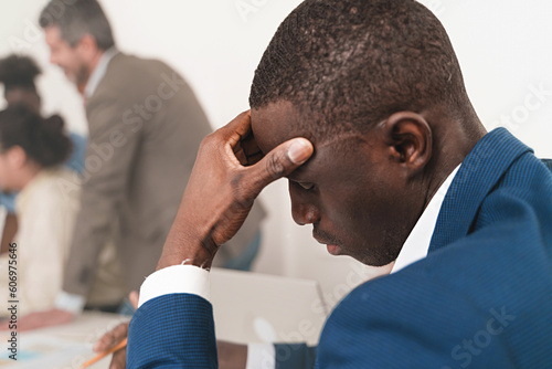 African businessman with headache in office - Stressed African man in formalwear holds his forehead in discomfort, blurred coworkers in background