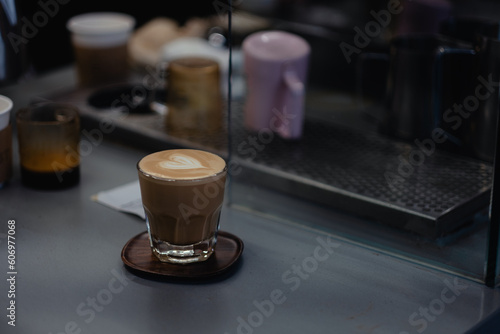 Busy scene at a café — a cappuccino is served with heart-shaped latte art (coffee foam art) at a coffee bar, with an espresso machine and other barista equipment in the background.
